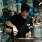 Young person cooking on a griddle in a bustling kitchen.