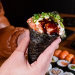 サーモンとねぎを巻いた手巻き寿司を手に持つクローズアップ、奥に巻き寿司の盛り合わせが並ぶ