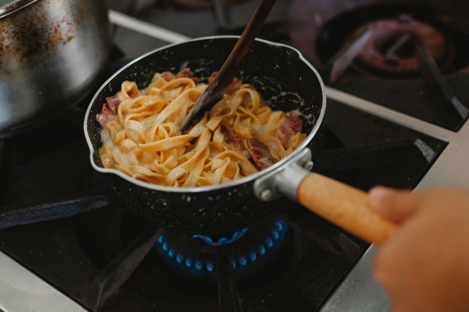 フライパンでベーコン入りのクリームパスタを木べらで炒め合わせている様子