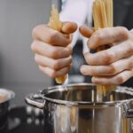両手で乾燥パスタをつかみ、湯気の立つ鍋に入れようとしている様子