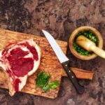 木のまな板に置かれた生の牛肉の塊と、包丁やハーブをすりつぶすすり鉢
