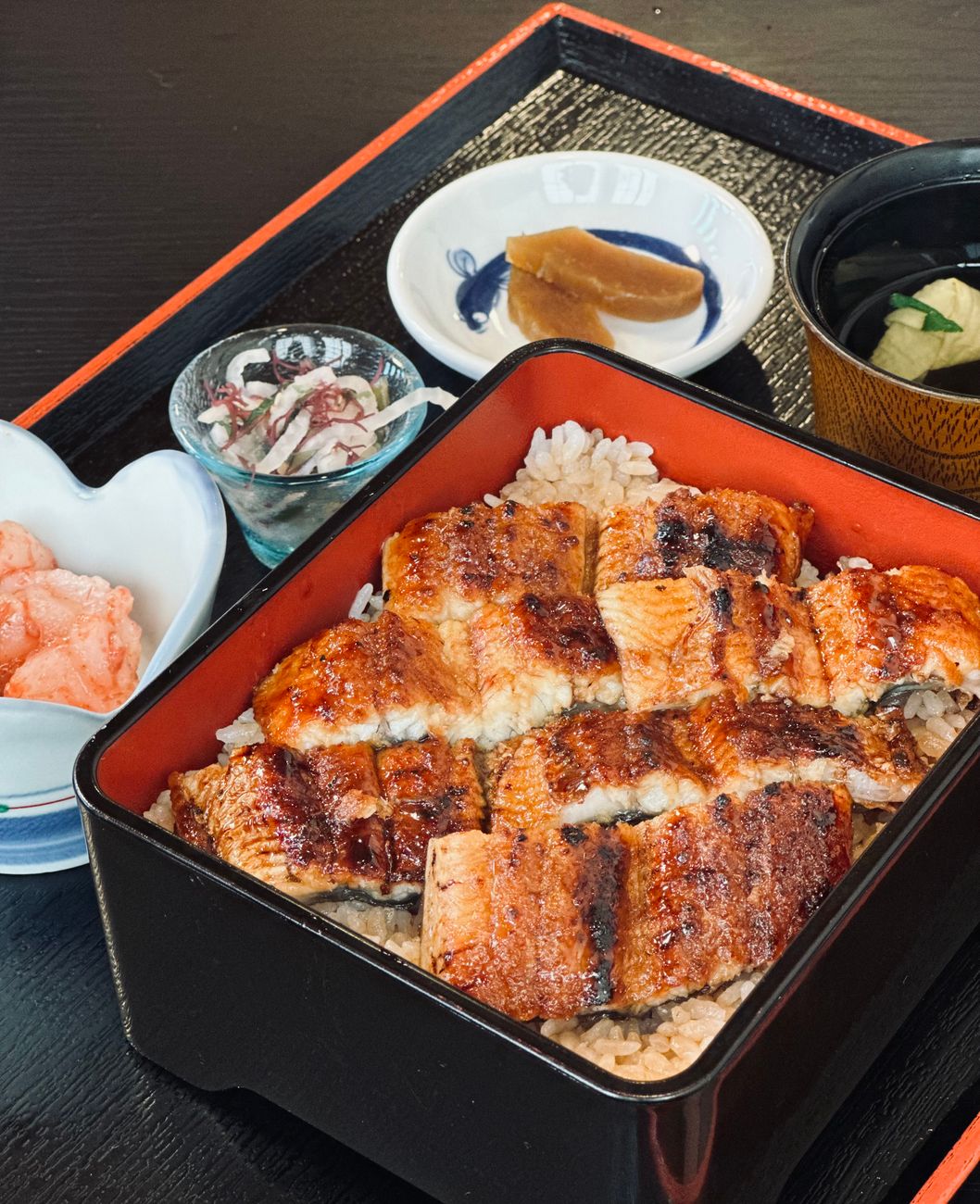 朱塗りの重箱にご飯を詰め、香ばしく焼き上げた鰻の蒲焼きを並べたうな重。奈良漬けや紅生姜、お吸い物が添えられている