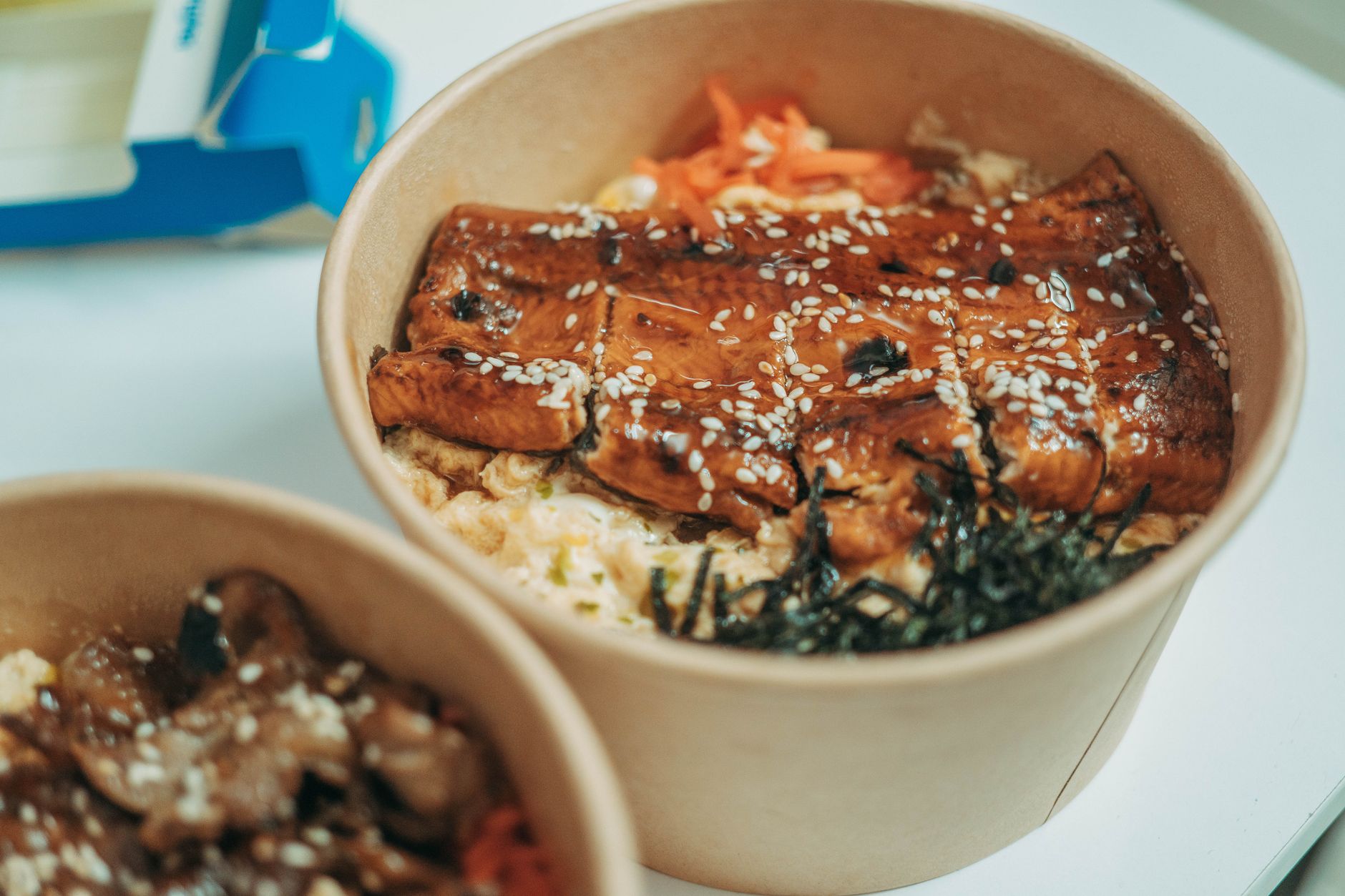紙製のテイクアウト容器に入った鰻丼。刻み海苔やにんじんの甘酢漬け、白ごまがトッピングされている