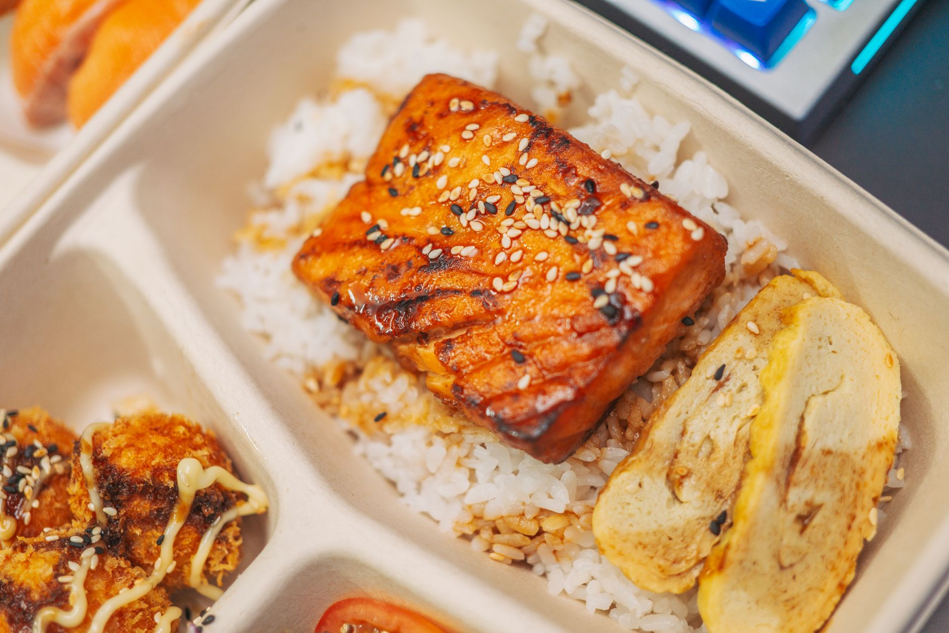 テイクアウト容器の中で、ごま入りの照り焼きだれをまとった魚の切り身がご飯にのっている。卵焼きやかつサンドも一緒に詰められている